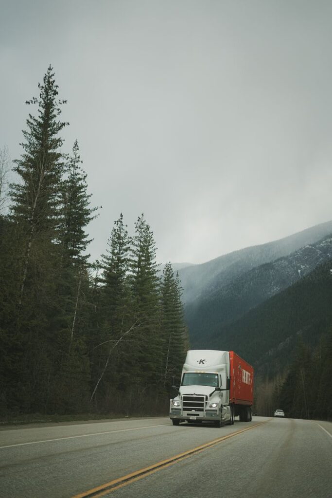A delivery truck drives along a scenic highway surrounded by mountains and evergreen trees.